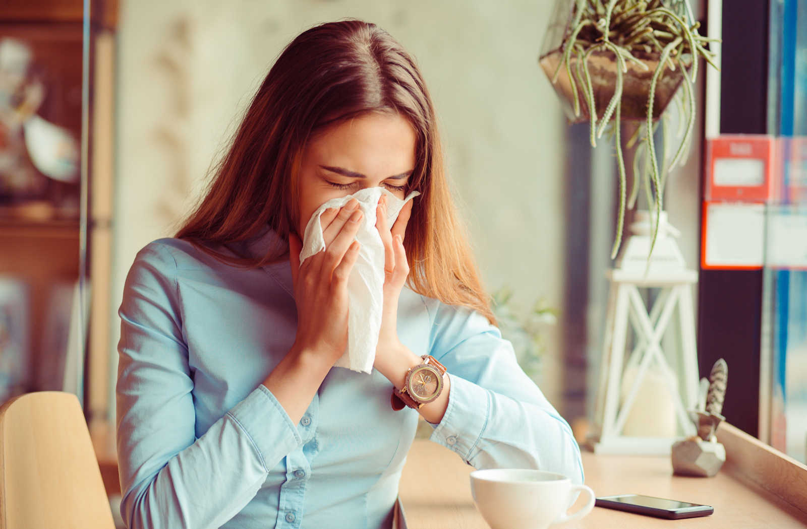 A young woman sitting at a table with a cup of tea. She is sneezing and holding a tissue with both hands to cover her nose and mouth.